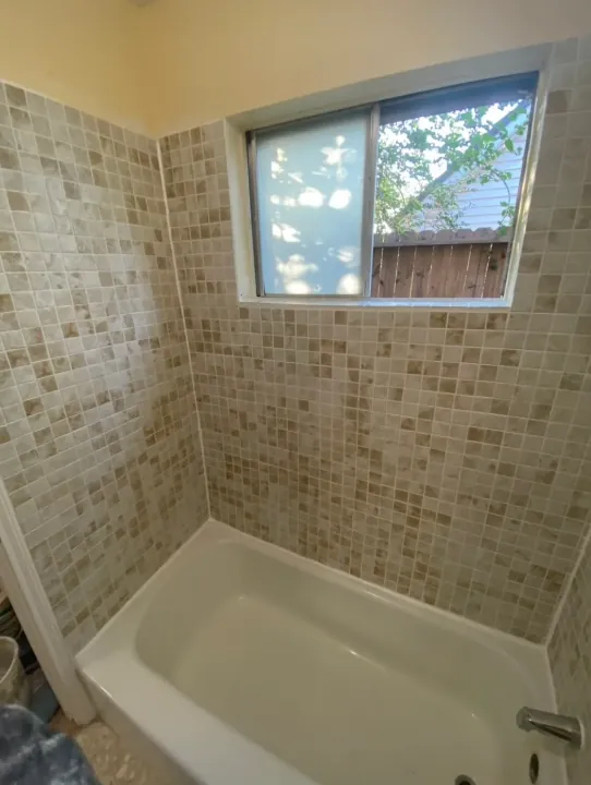 Tub surround with beige mosaic-style wall tile, a window, and a white tub